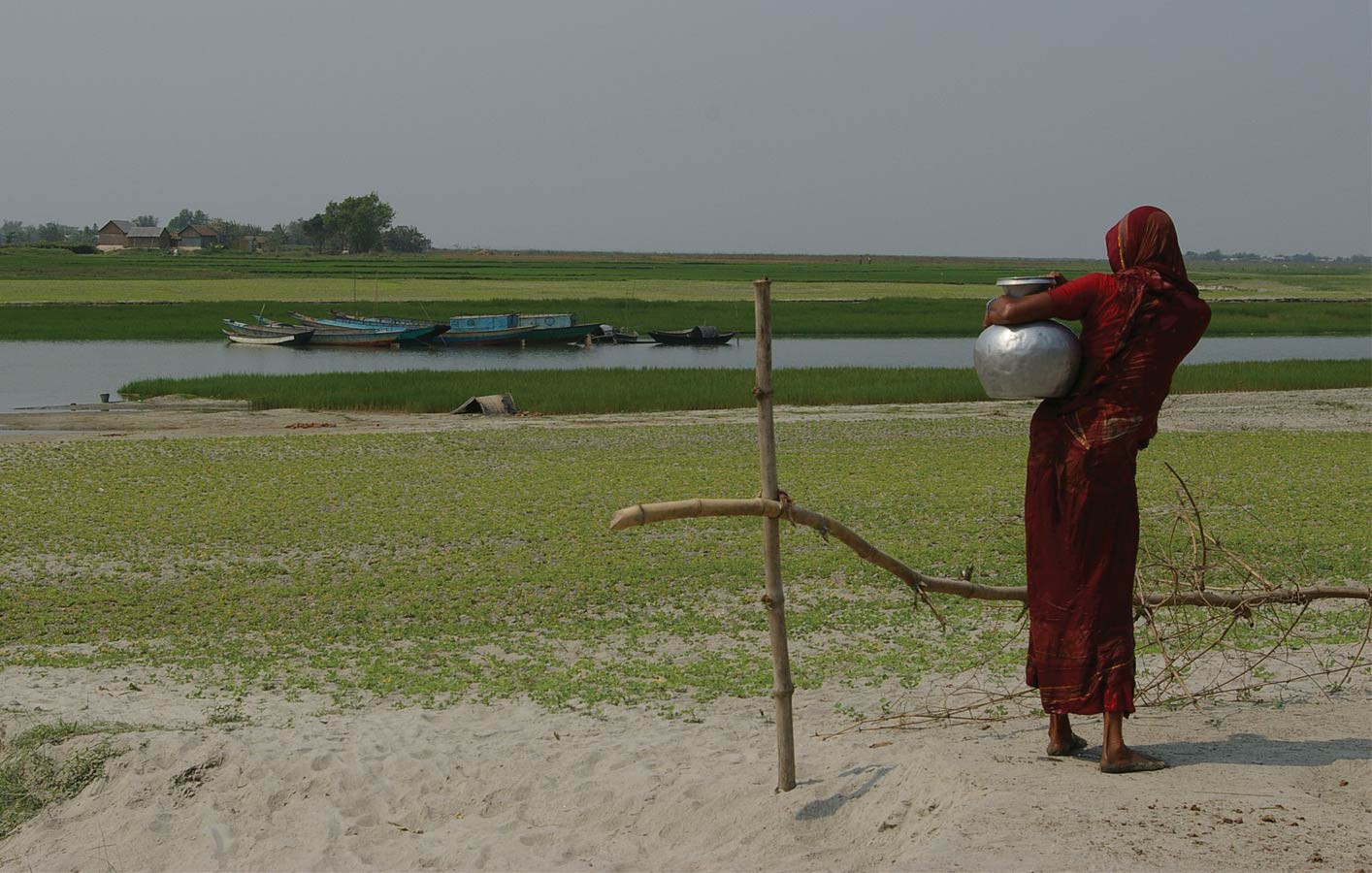 <p>A woman carrying water stands before a shallow waterway separating char settlements. There are typically no roads or formal access to chars.</p>
